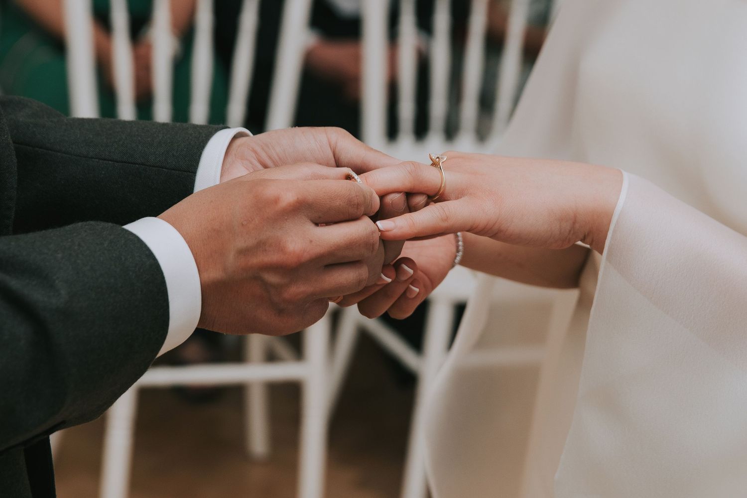 Groom placing a wedding ring on brides finger during a ceremony, with white chairs visible in the background.
