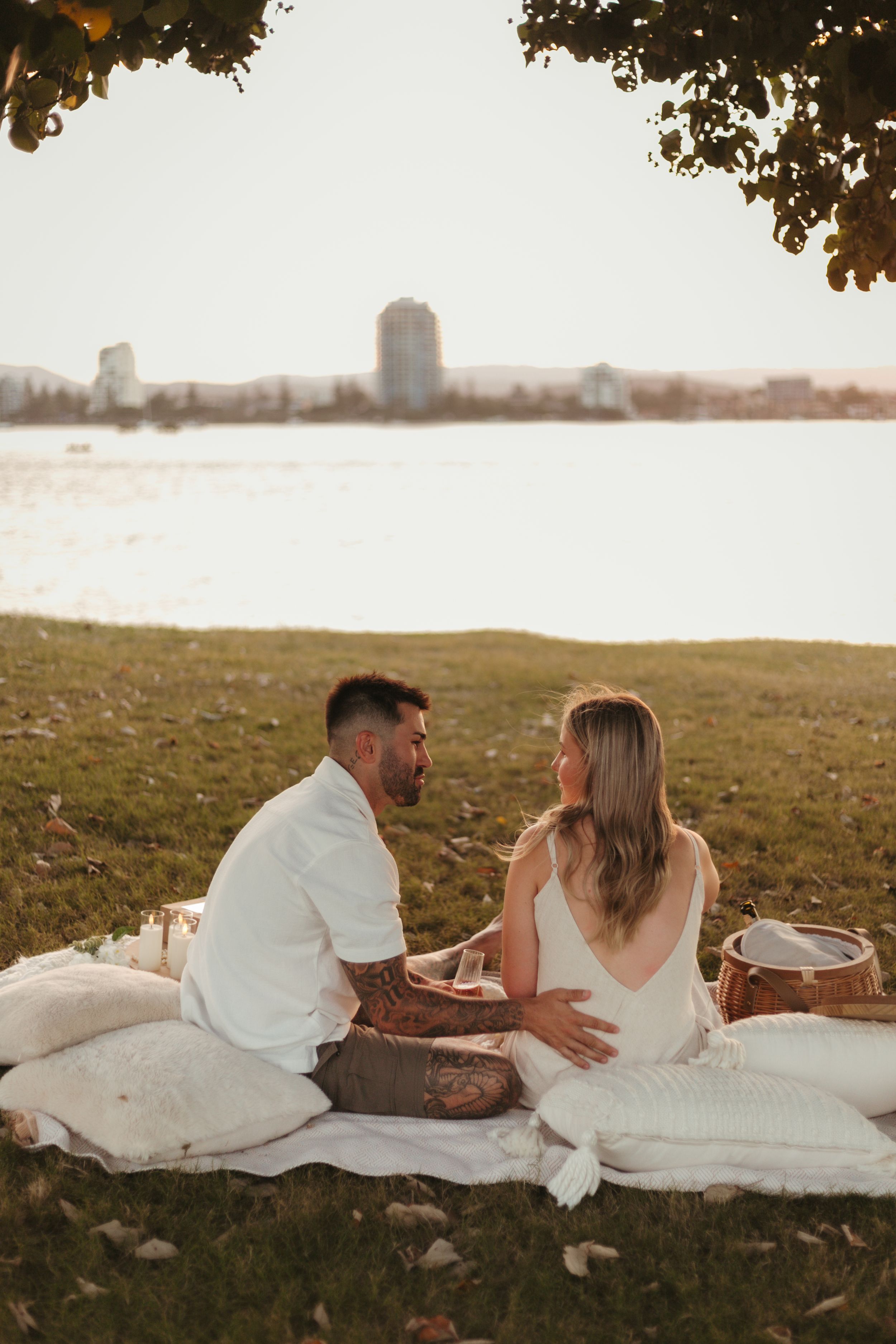 Couple sitting on a white blanket by the waterfront at sunset, sharing a romantic picnic outdoors.
