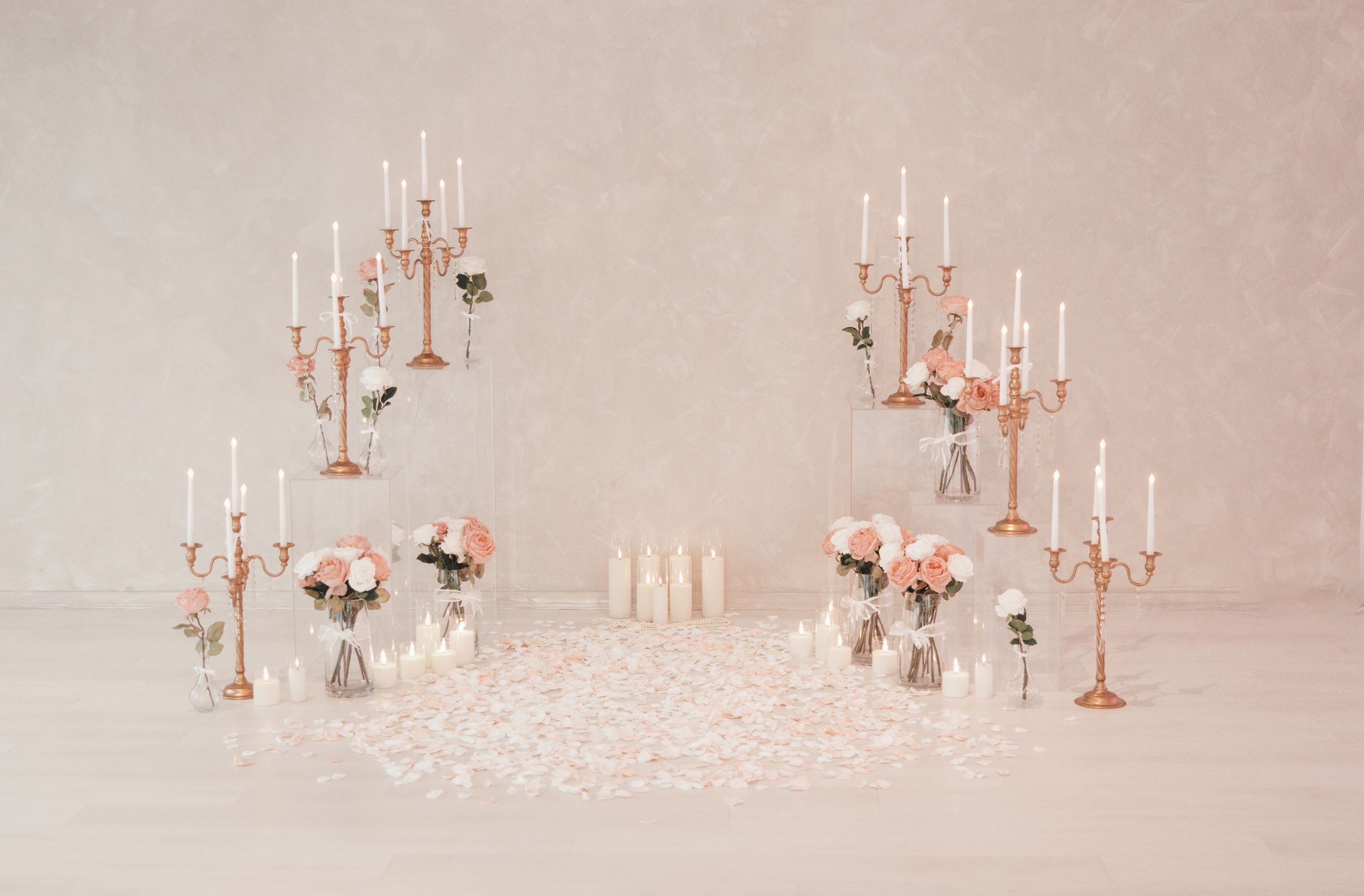 Elegant wedding ceremony backdrop with gold candelabras, white candles, blush roses, and rose petals on a white floor.