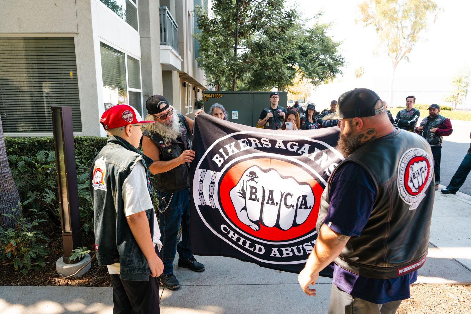 Bikers Against Child Abuse members hold banner at outdoor rally near building.