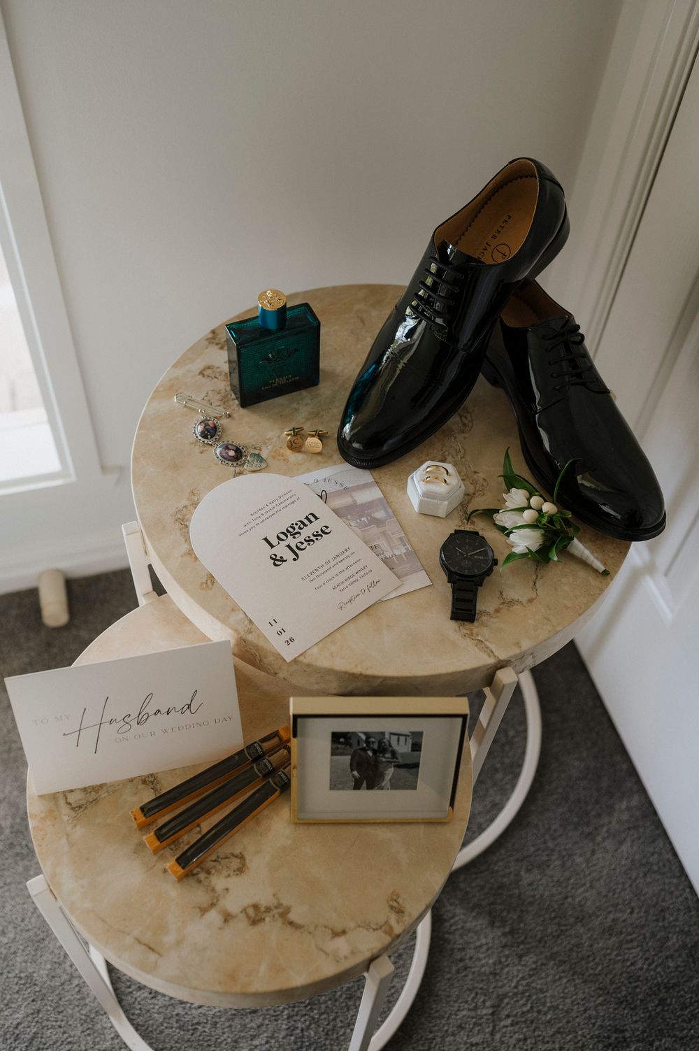 Groom wedding flat lay with black patent shoes, watch, boutonniere, invitation, cologne, and framed photo on marble tables.