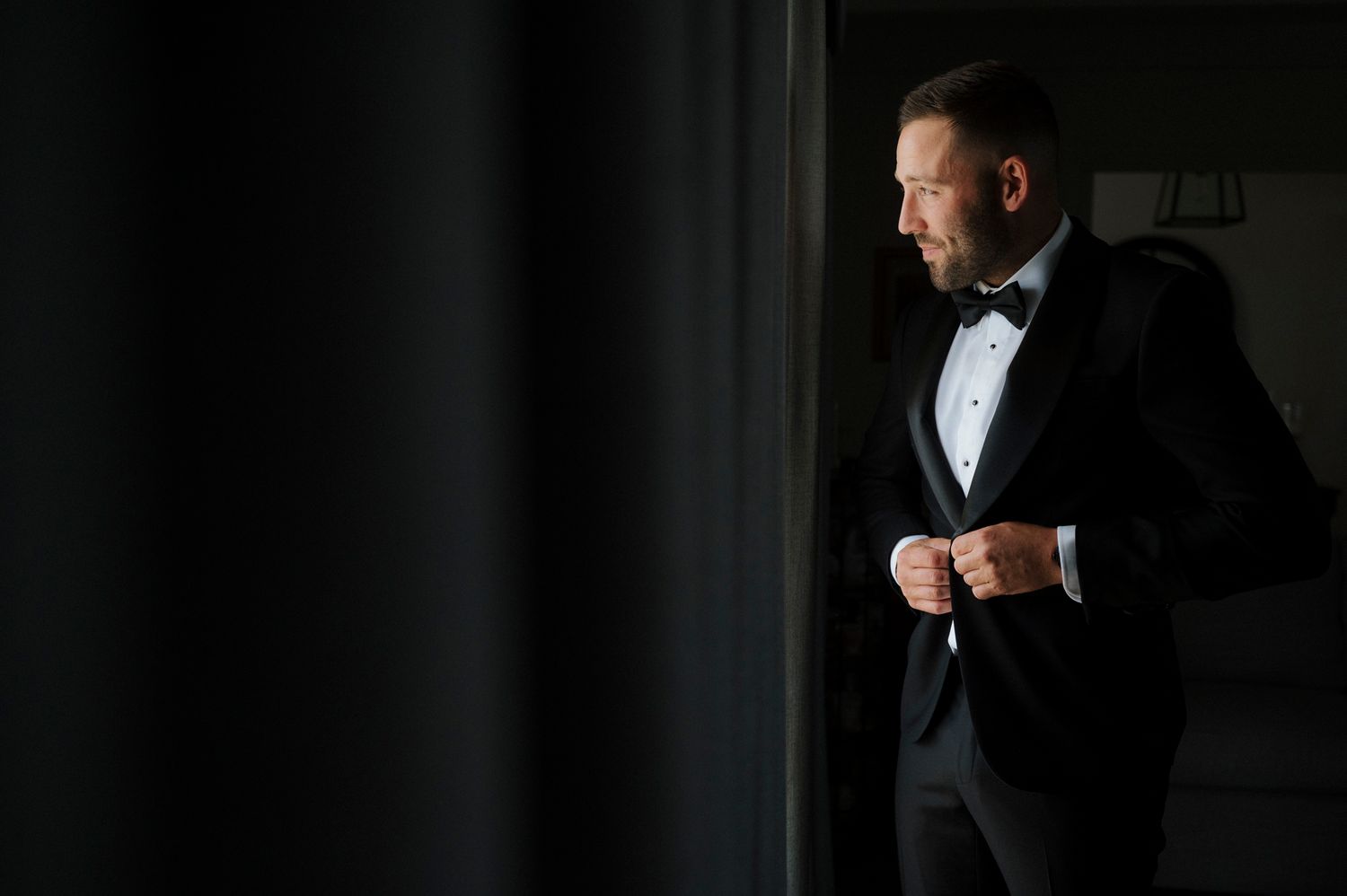 Elegant man in black tuxedo and bow tie buttoning jacket near window in dark room.