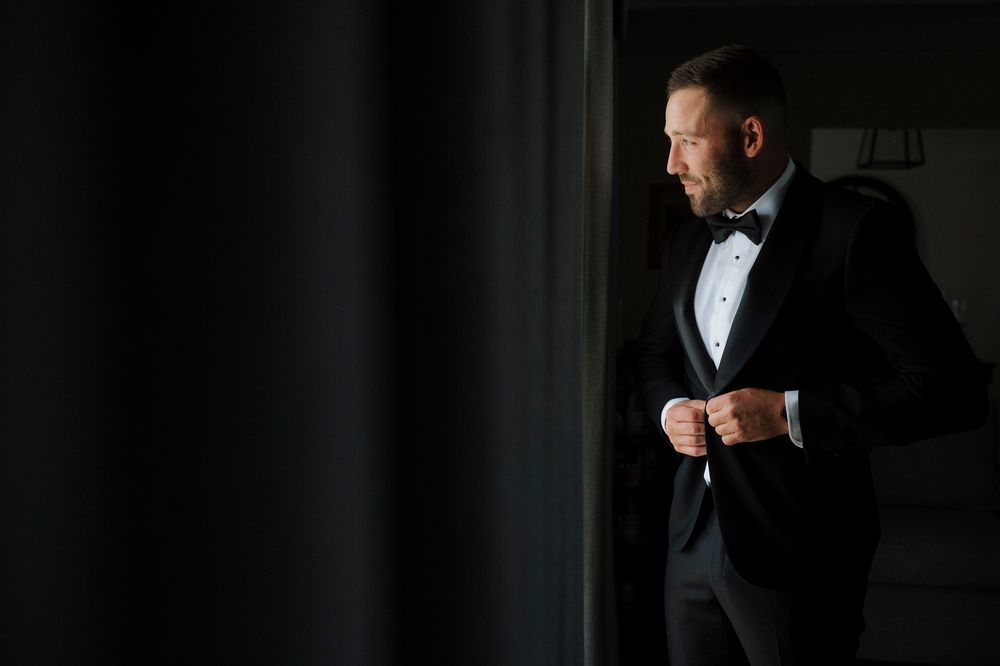 Elegant man in black tuxedo and bow tie buttoning jacket near window in dark room.