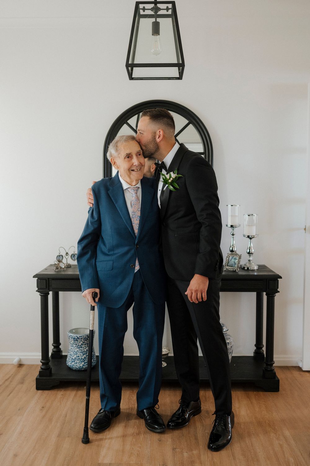 Groom in black tuxedo kisses elderly man in blue suit holding cane at wedding celebration.