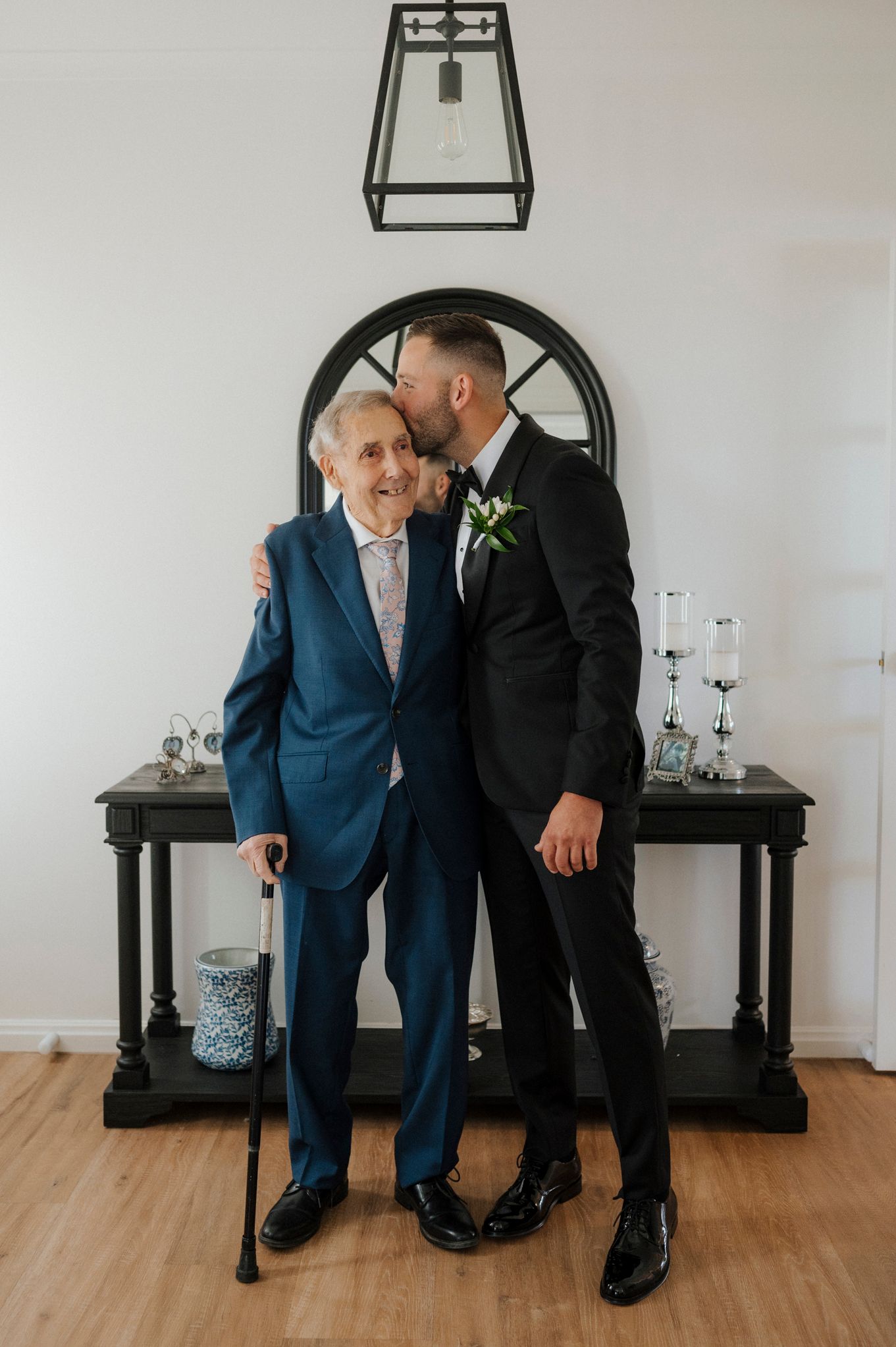 Groom in black tuxedo kisses elderly man in blue suit holding cane at wedding celebration.