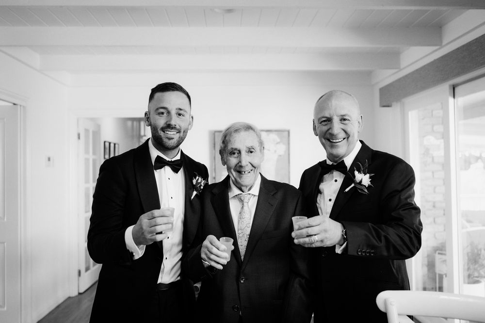 Three men in tuxedos smile and raise glasses in a black and white wedding photo.