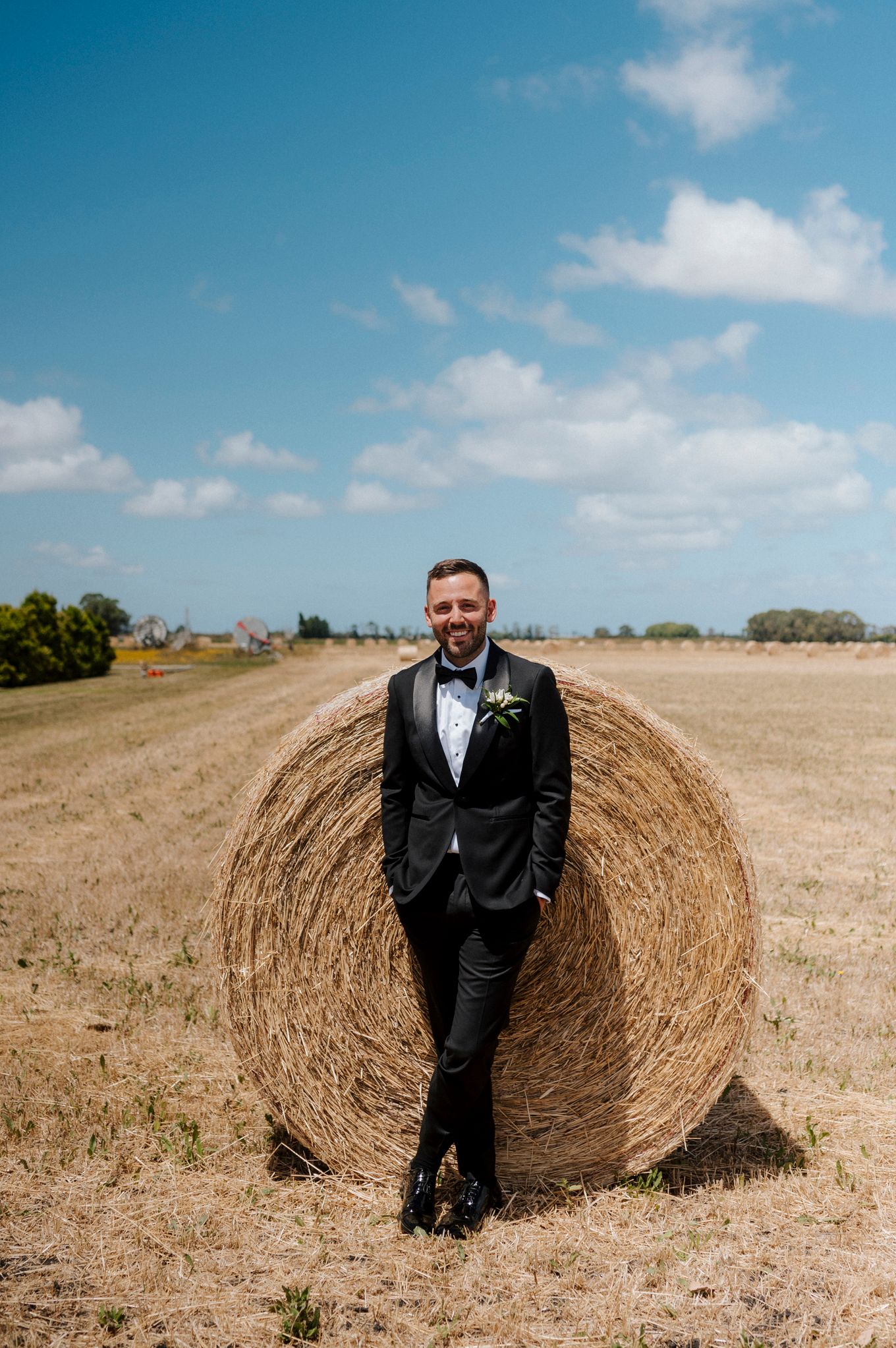 Groom in black tuxedo leaning against a round hay bale in a sunny field, smiling with a floral boutonniere.
