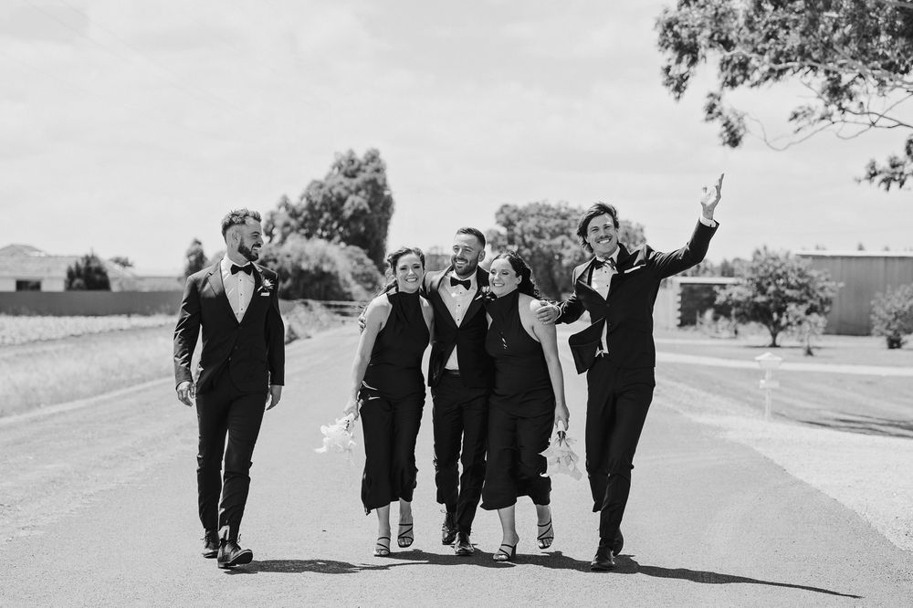 Black and white photo of a joyful wedding party in formal attire walking down a sunny road, arms raised in celebration.