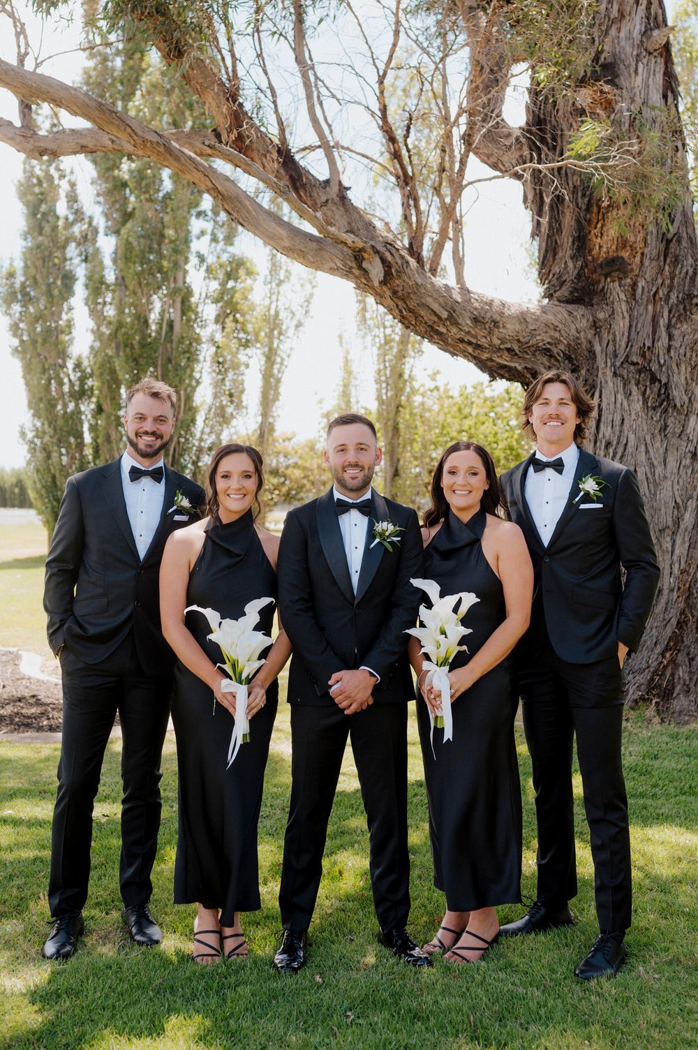 Wedding party in black formal attire posing outdoors under a large tree, bridesmaids holding white lily bouquets.