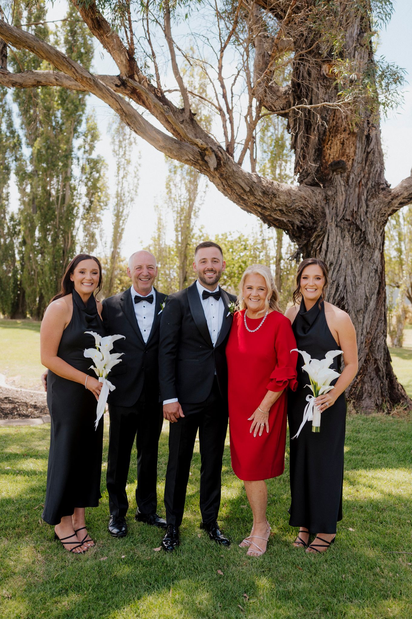 Family poses outdoors in formal attire near a large tree, with two women holding white floral bouquets at a wedding.