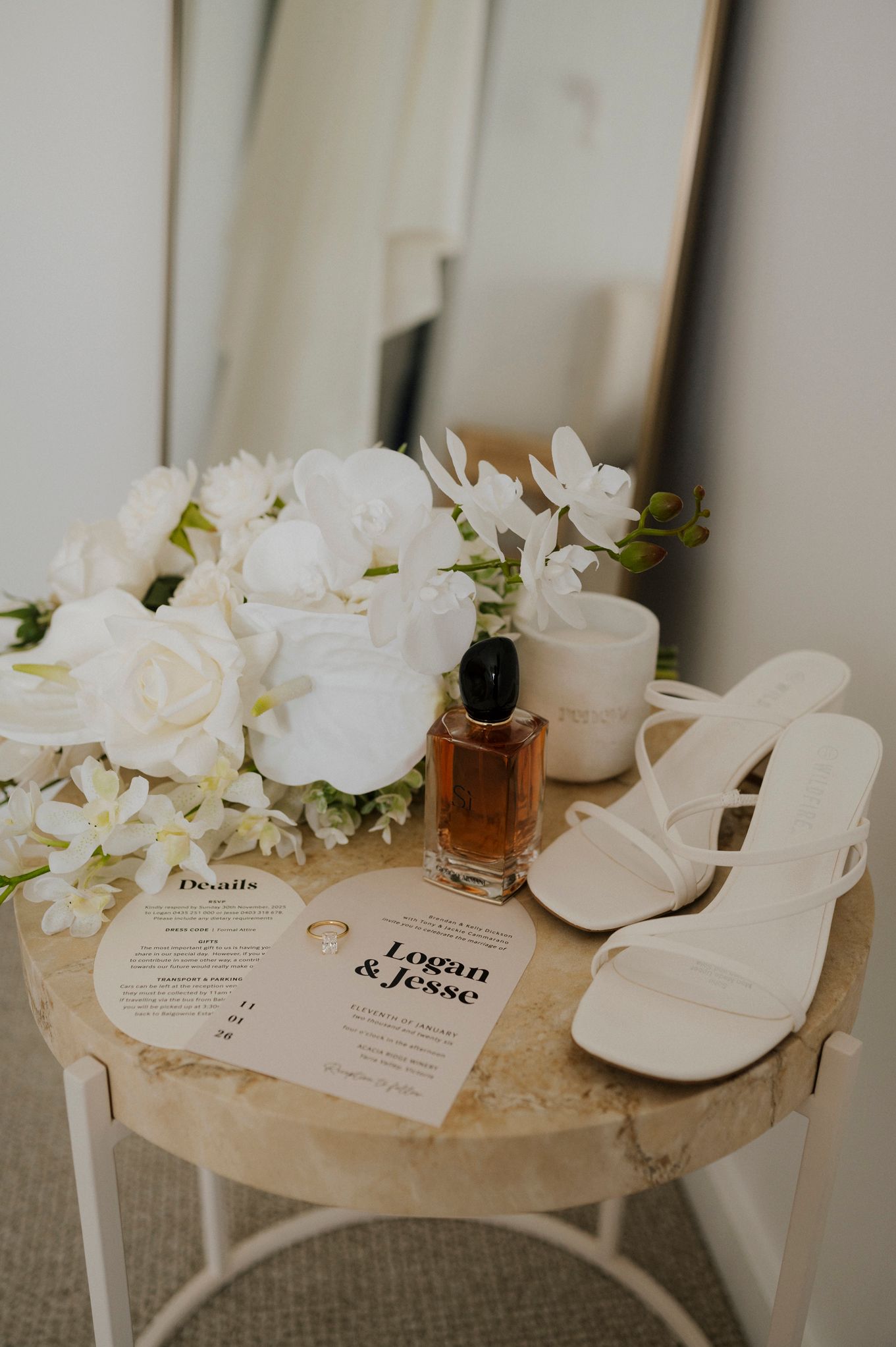 Elegant wedding flat lay with white heels, bouquet, perfume, and Logan & Jesse invitation cards on a wooden table.