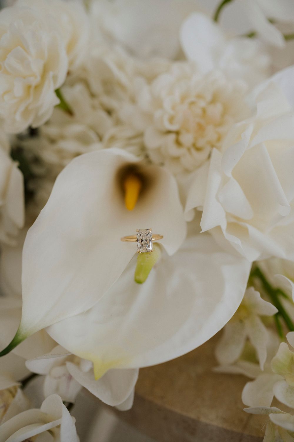 Gold diamond engagement ring resting on a white calla lily surrounded by cream roses and flowers.