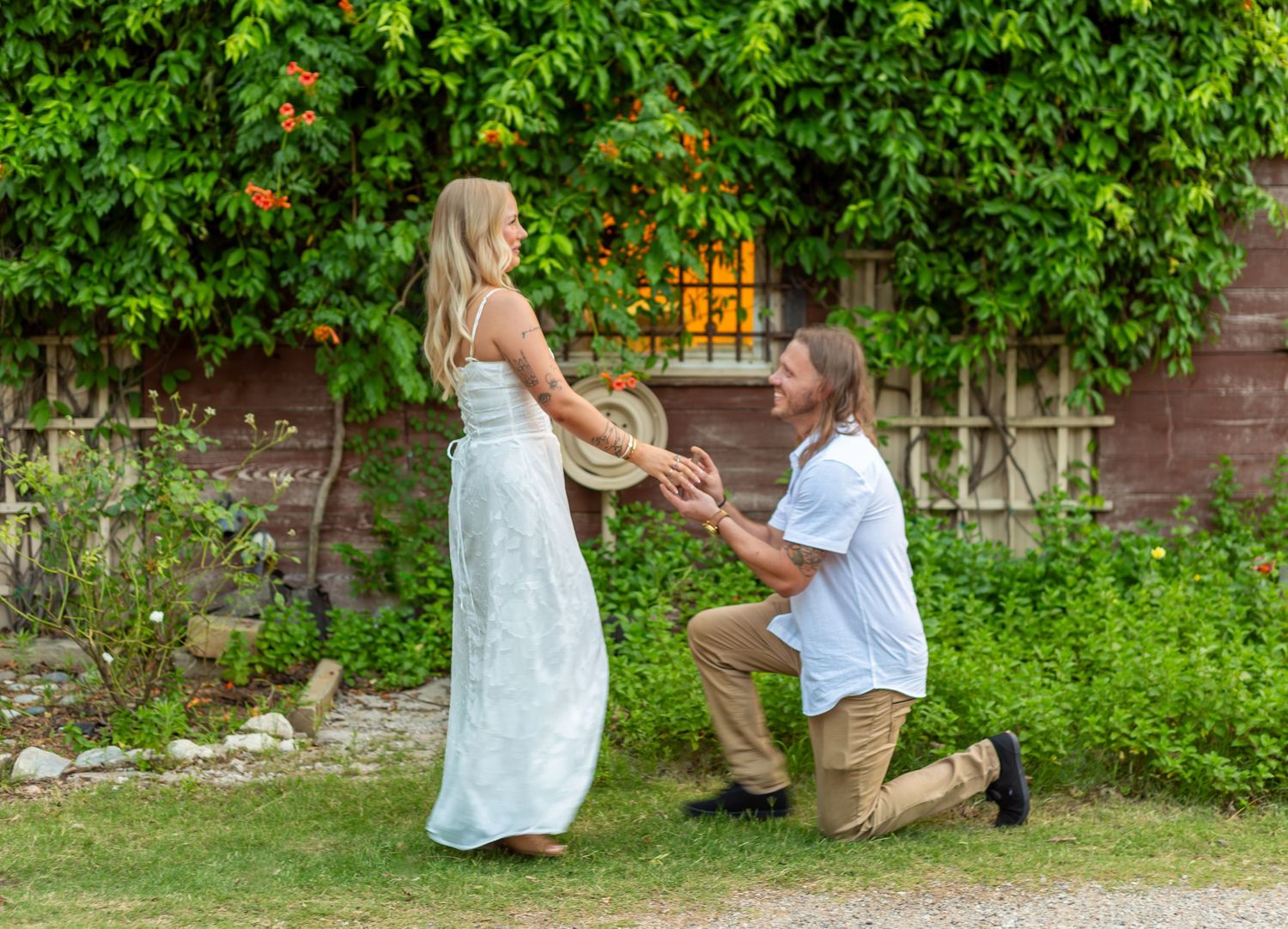 Man proposing on one knee to woman in white dress outdoors near brick building with lush green ivy backdrop.