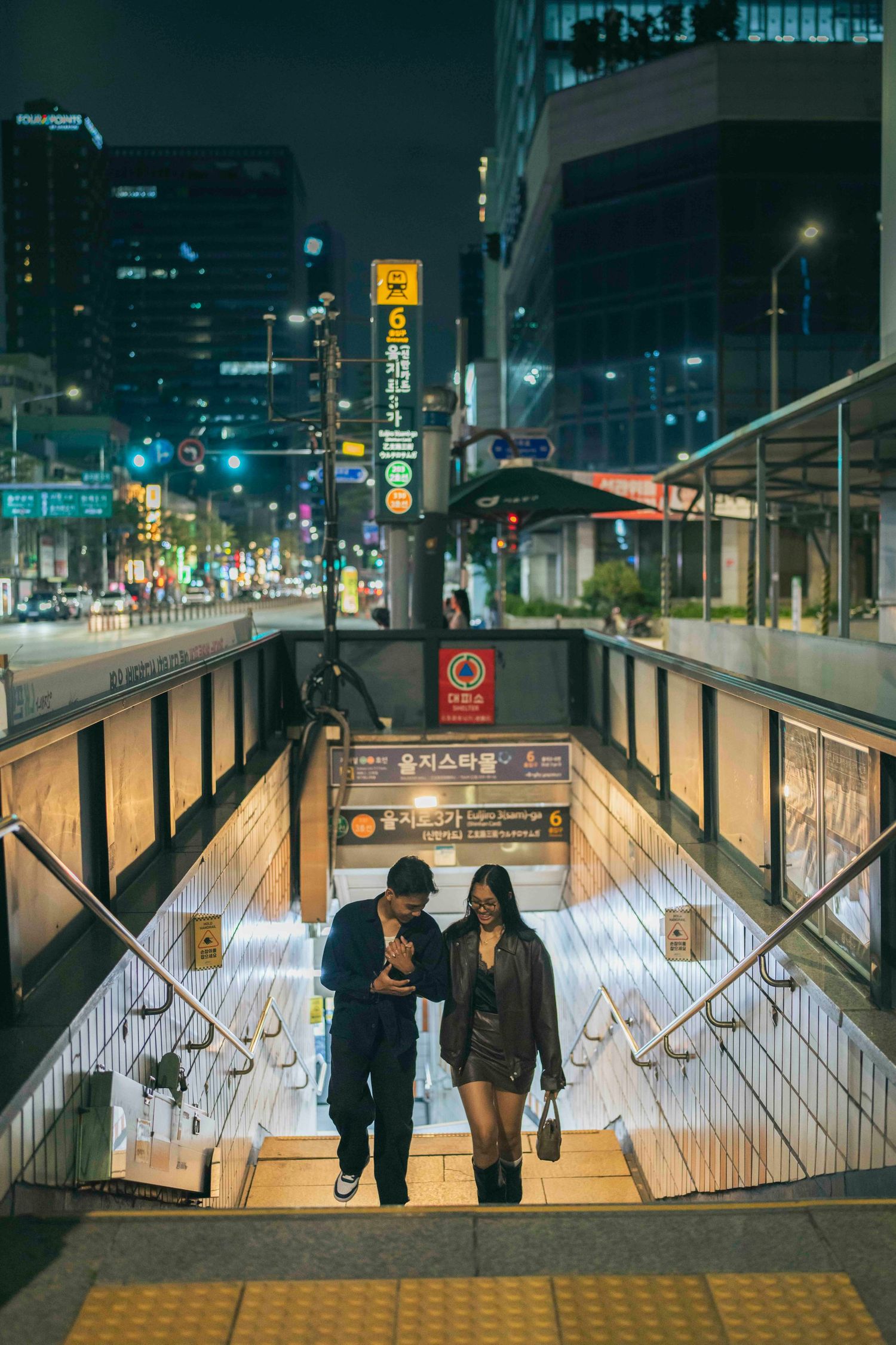 Couple emerging from Seoul subway station at night, city lights glowing behind them on a busy urban street.