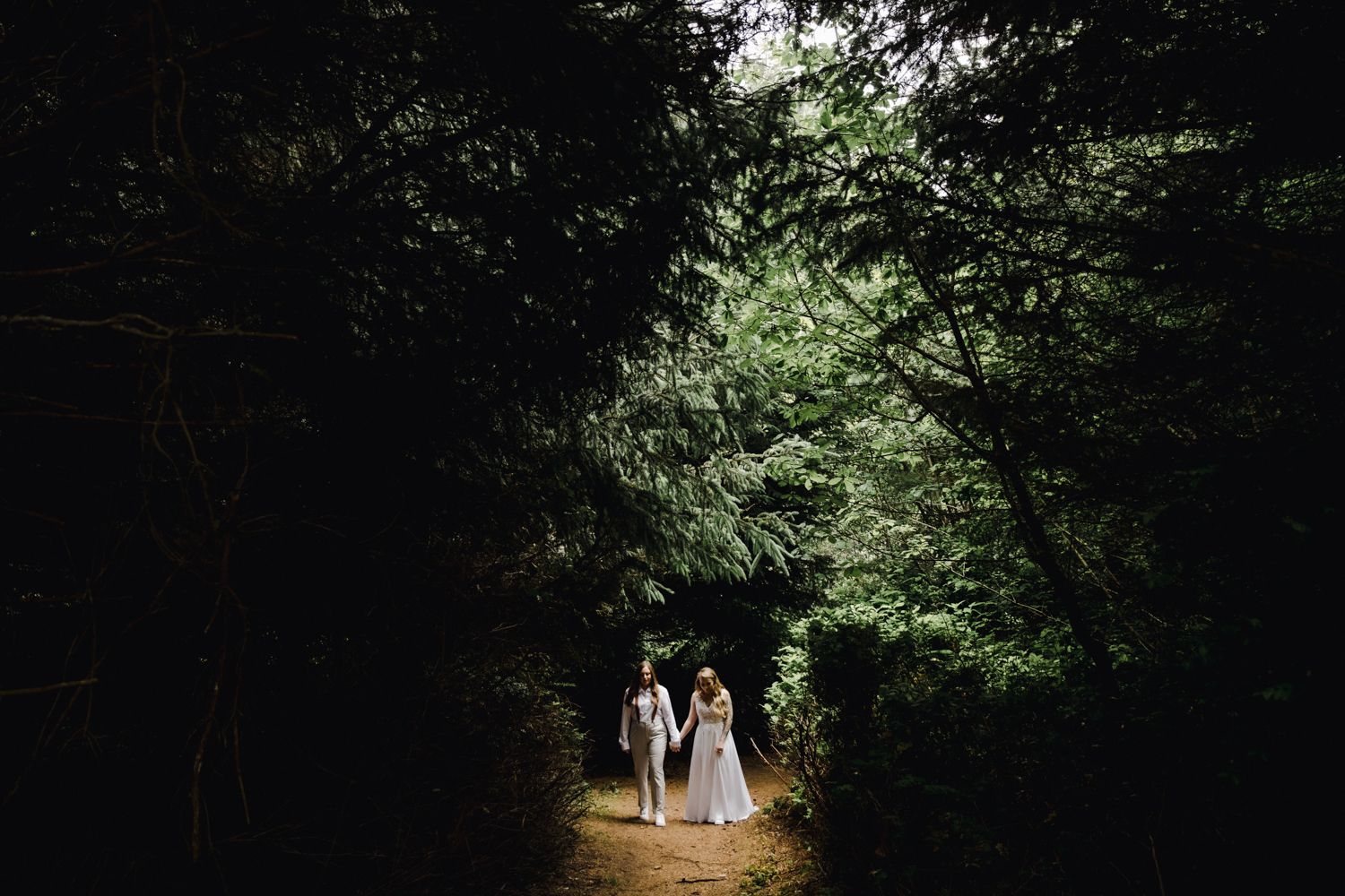Bride and groom walking hand in hand through a lush green forest tunnel on their wedding day.