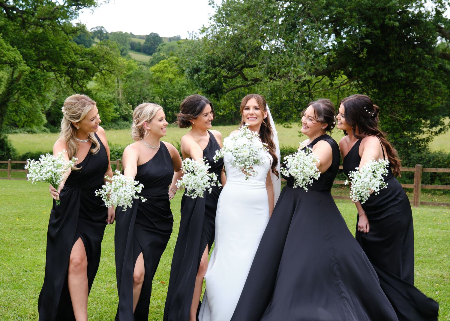 Bride in white gown with five bridesmaids in black dresses holding white bouquets laughing outdoors on green lawn. White hart inn wedding