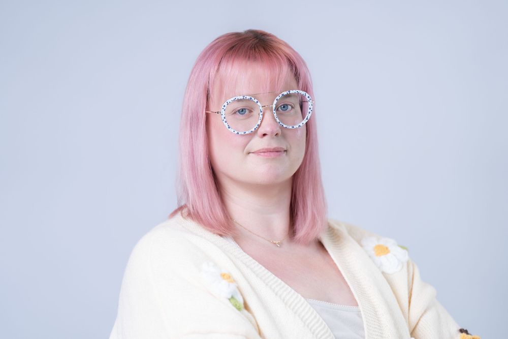 Young woman with pink hair and decorative round glasses wearing a cream cardigan against a gray background.