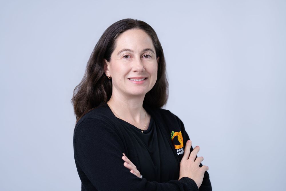 Professional woman with crossed arms wearing a black sweatshirt, smiling against a light gray background.