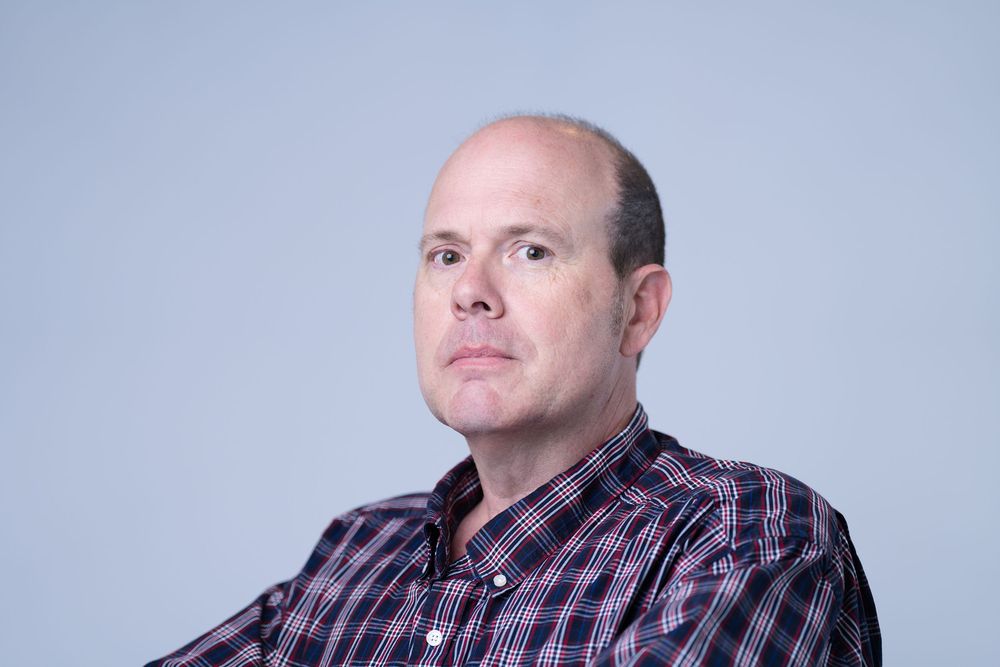 Middle-aged bald man in plaid shirt poses for professional headshot against light gray background.
