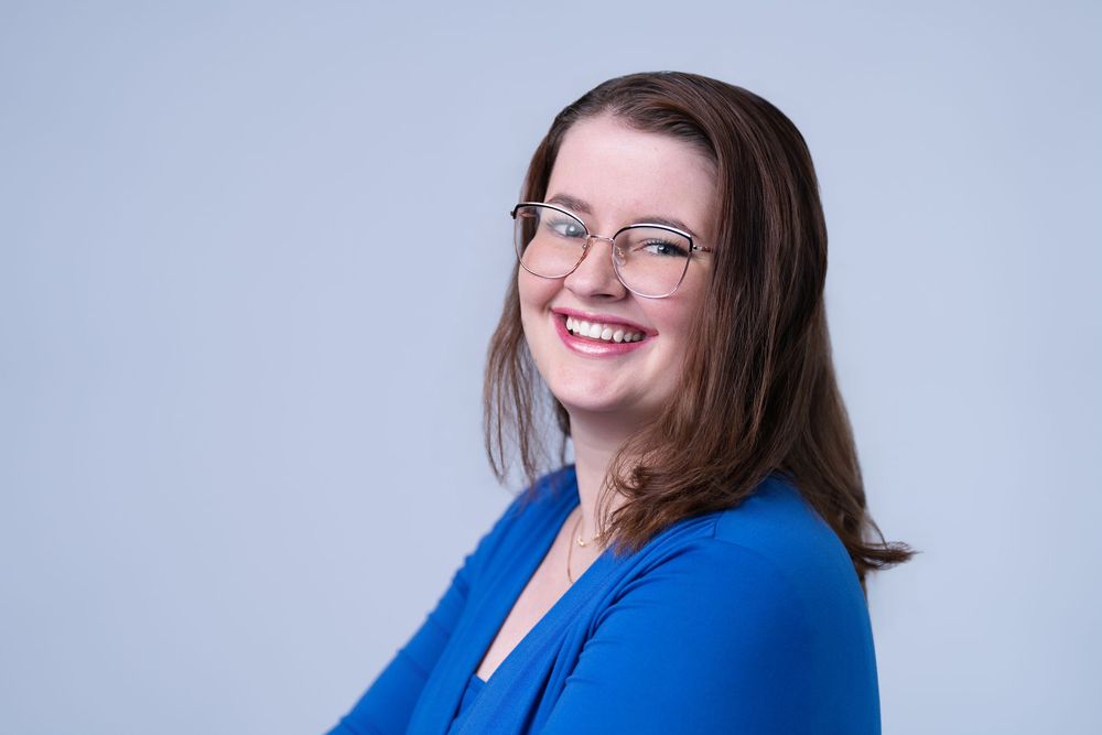 Smiling woman with glasses and brown hair wearing a blue blazer poses against a light gray background.