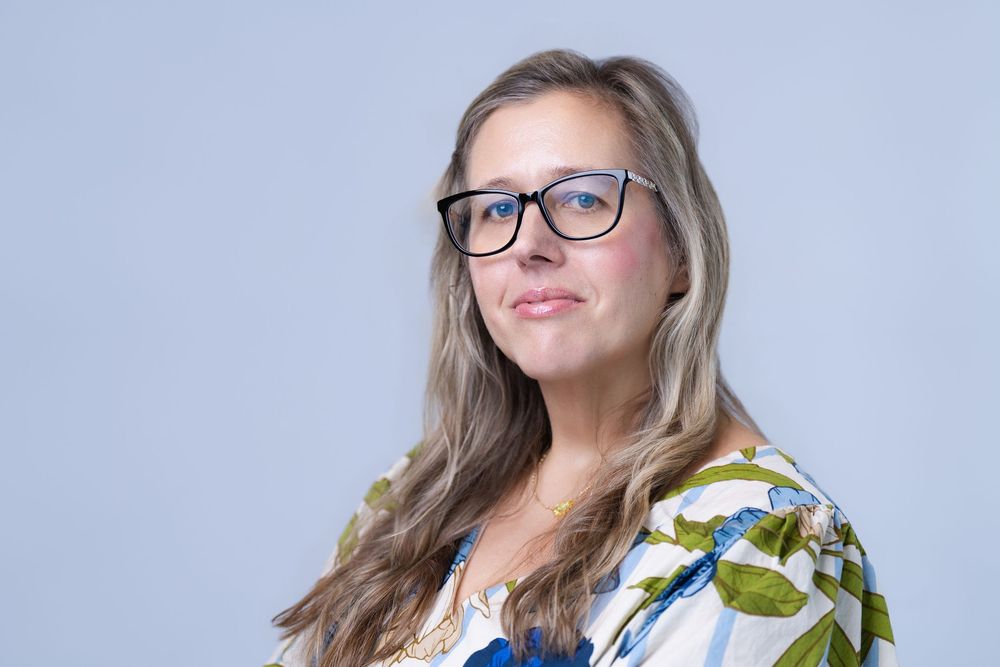 Woman with long blonde hair and black glasses wearing a floral blouse, posing against a light gray background.