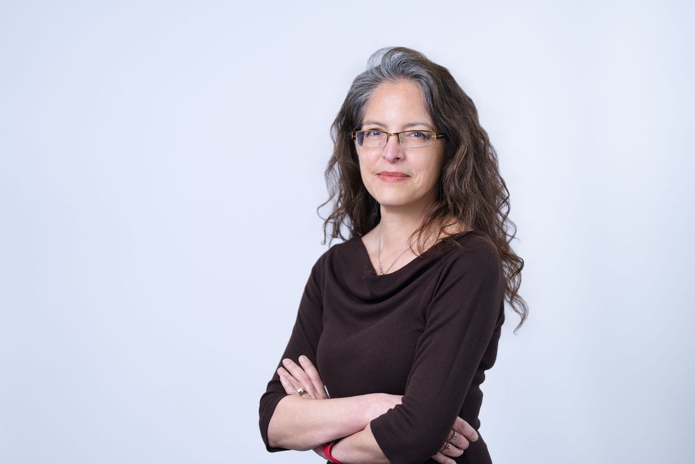Professional woman with curly hair and glasses, arms crossed, wearing a brown top against a light background.