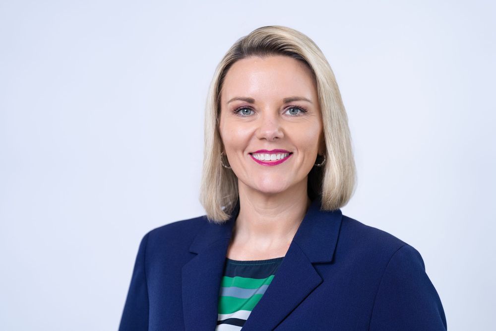 Professional headshot of a smiling blonde woman in a navy blazer and striped top against a light background.