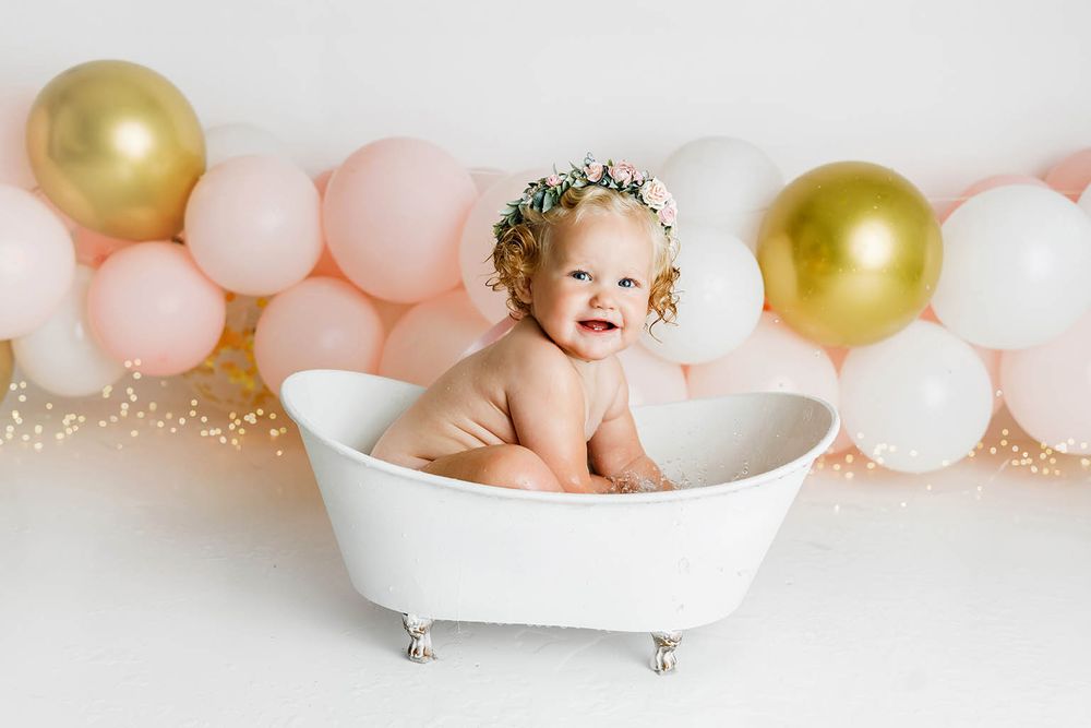 Adorable baby girl in floral crown sitting in white mini bathtub with pink and gold balloons backdrop.