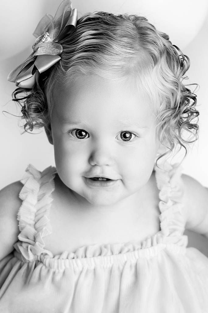 Black and white portrait of a curly haired baby girl with a bow wearing a ruffled dress, smiling softly.