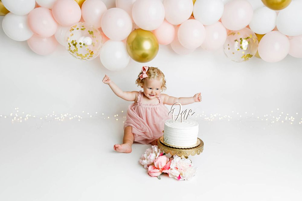 Baby girl in pink romper reaches toward white cake during first birthday cake smash with pink and gold balloon arch.