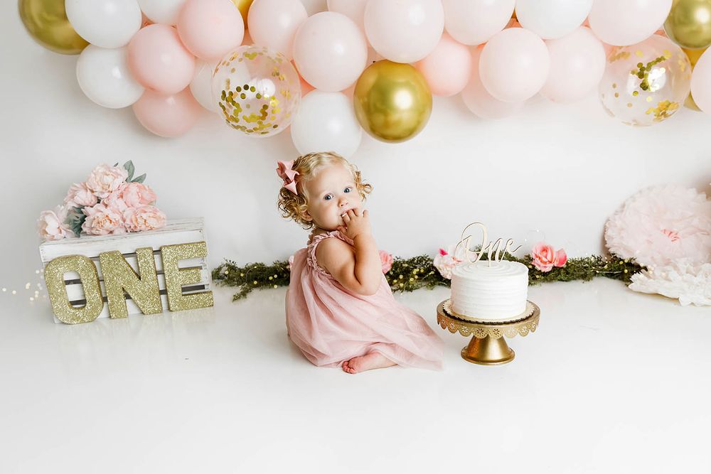 Baby girl in pink dress sits beside white cake with ONE sign and pink gold balloon arch for first birthday cake smash.