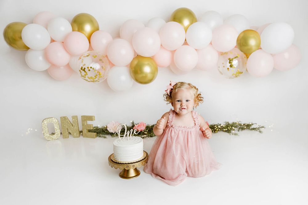 Baby girl in pink dress celebrates first birthday with white cake and ONE sign, pink and gold balloon garland backdrop.