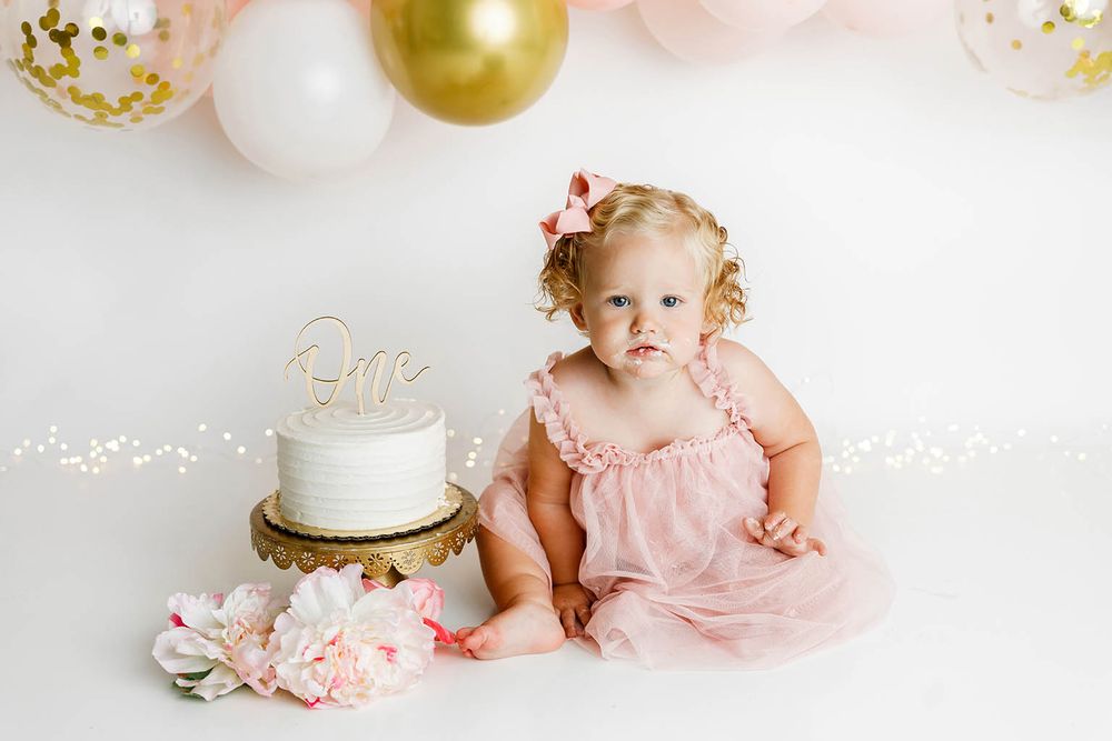 Baby girl in pink dress sits by white cake with gold topper, surrounded by pink and gold balloons for first birthday.