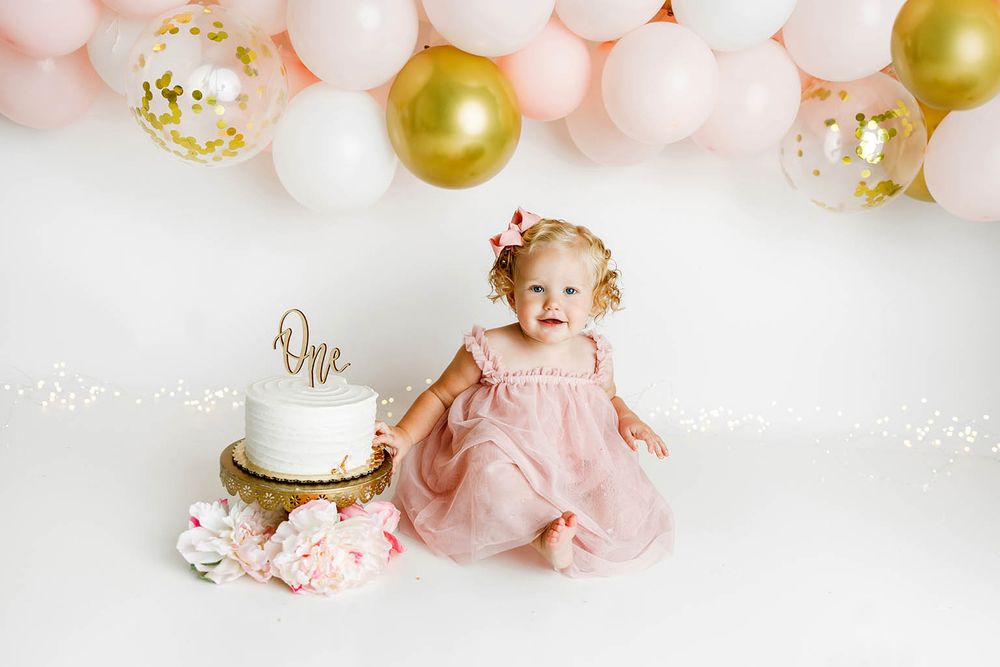 Adorable baby girl in pink dress sits next to white cake during first birthday cake smash with gold and pink balloons.
