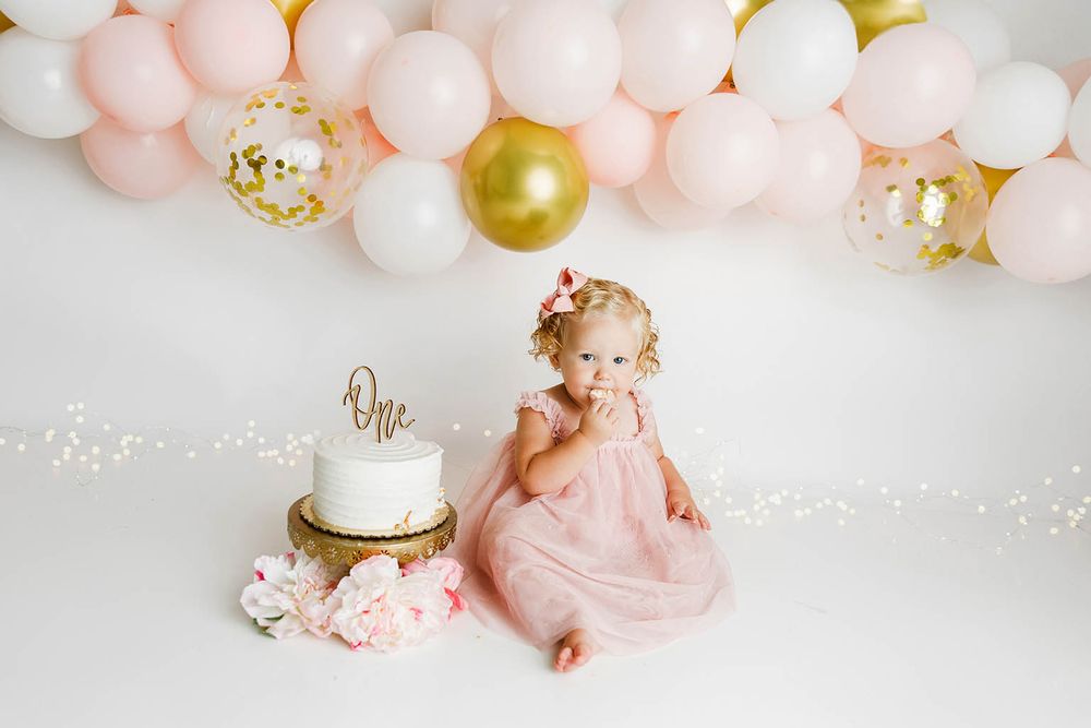 Adorable baby girl in pink tulle dress sits beside white One cake with pink gold balloon arch for first birthday smash.
