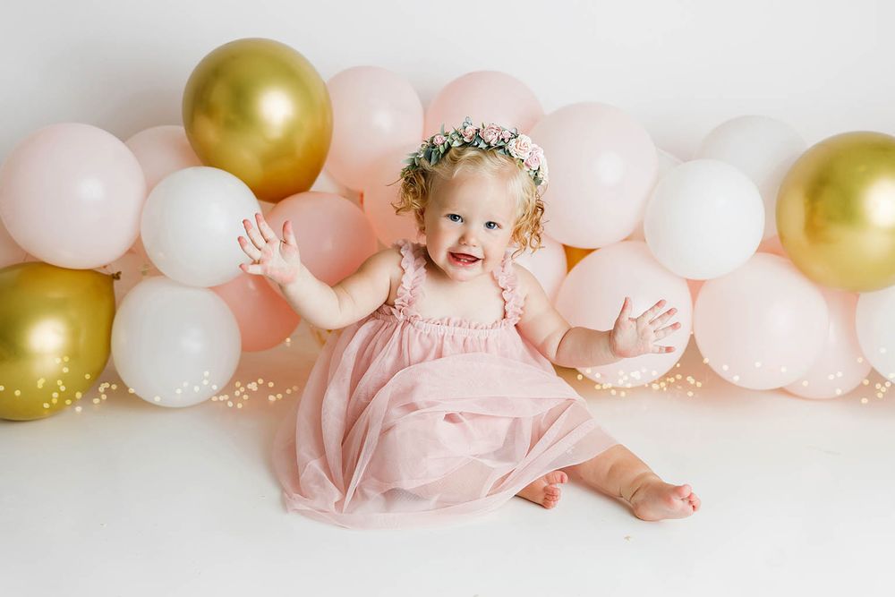 Baby girl in pink dress with floral crown sits smiling among pink, white, and gold balloons.