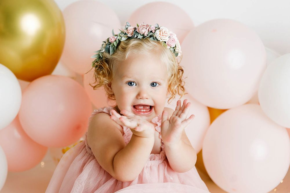 Smiling baby girl in pink dress and floral crown surrounded by pink and gold balloons at first birthday party.