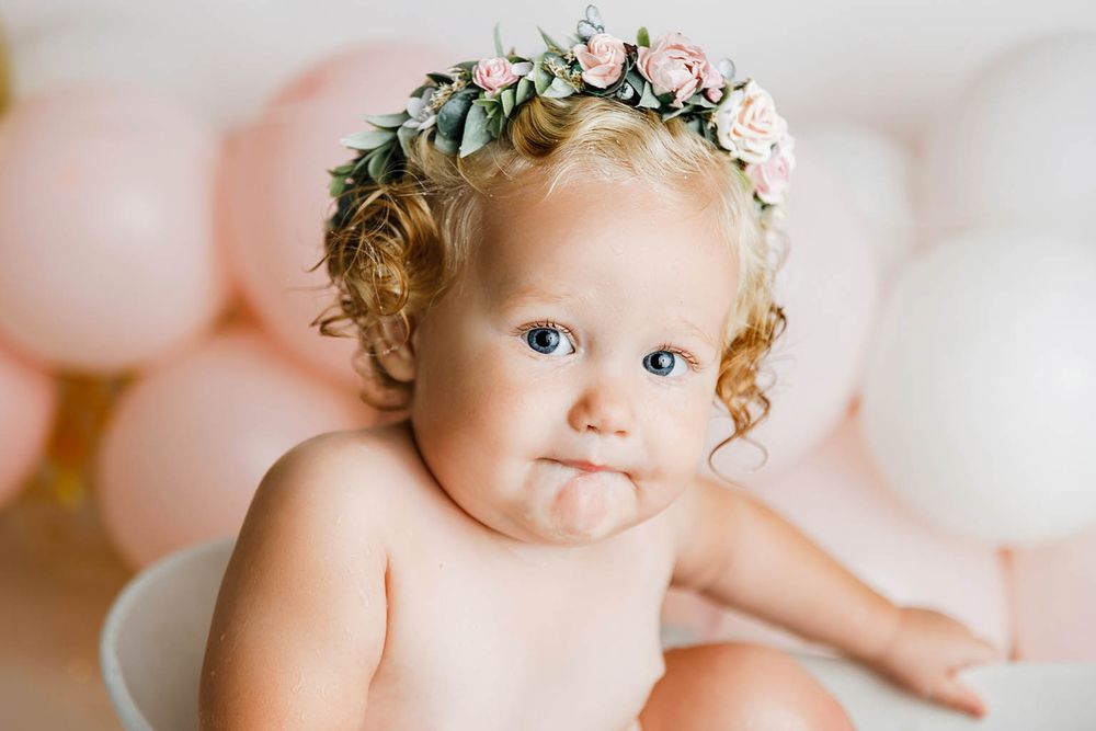 Adorable baby girl with curly hair wearing a floral crown surrounded by pink and white balloons.