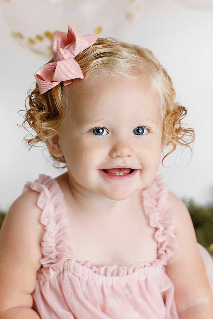 Cute toddler girl with blue eyes and curly blonde hair wearing pink bow and ruffled pink dress.