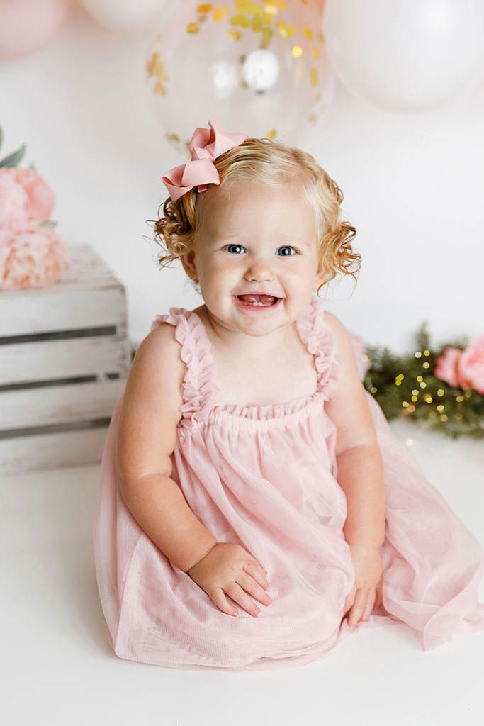 Smiling baby girl with curly blonde hair and pink bow wearing pink dress at first birthday cake smash photo session.