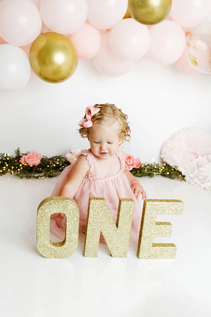 Baby girl in pink dress sitting next to gold glitter ONE letters with pink and gold balloon garland.