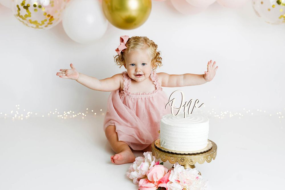 Baby girl in pink romper celebrating first birthday smash cake session with white cake and balloons.