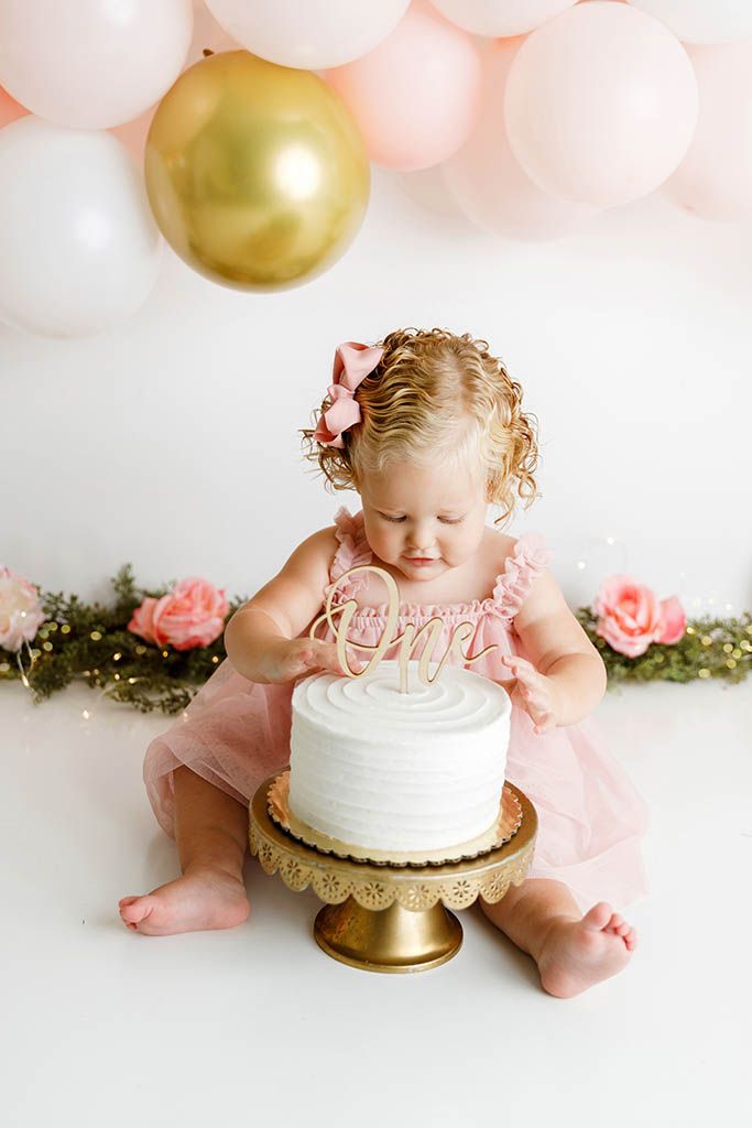 Baby girl in pink tutu smashes white cake on gold stand at first birthday cake smash photo session.
