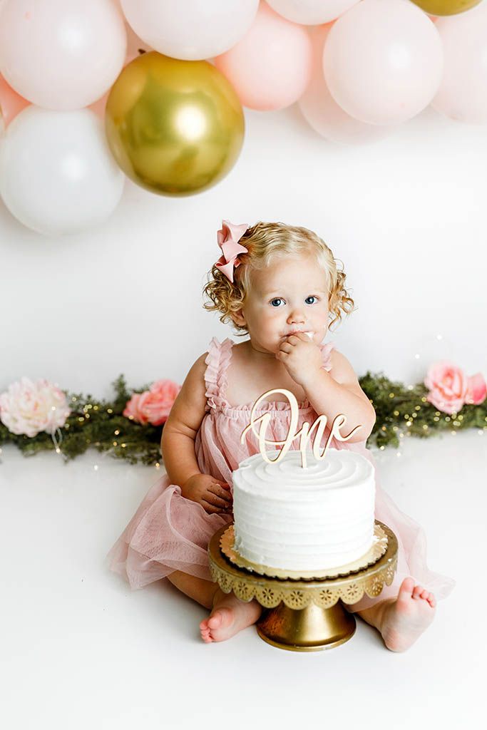 Baby girl in pink ruffled dress touches white frosted cake with One topper surrounded by pink gold balloons at first birthday.
