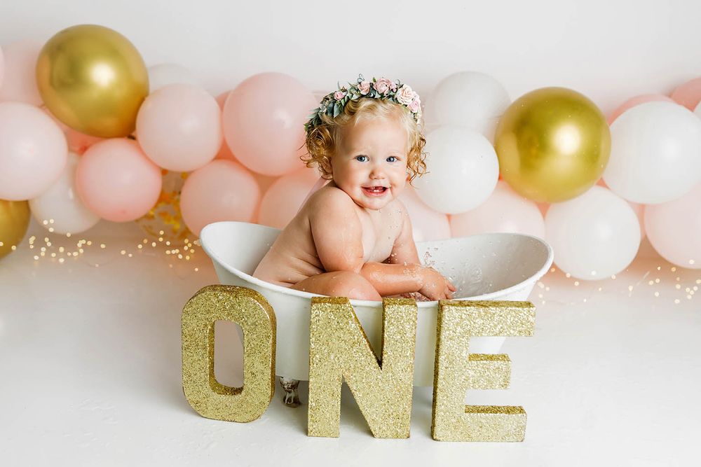 Smiling baby girl sits in white tub beside gold glitter ONE sign at first birthday photo session.