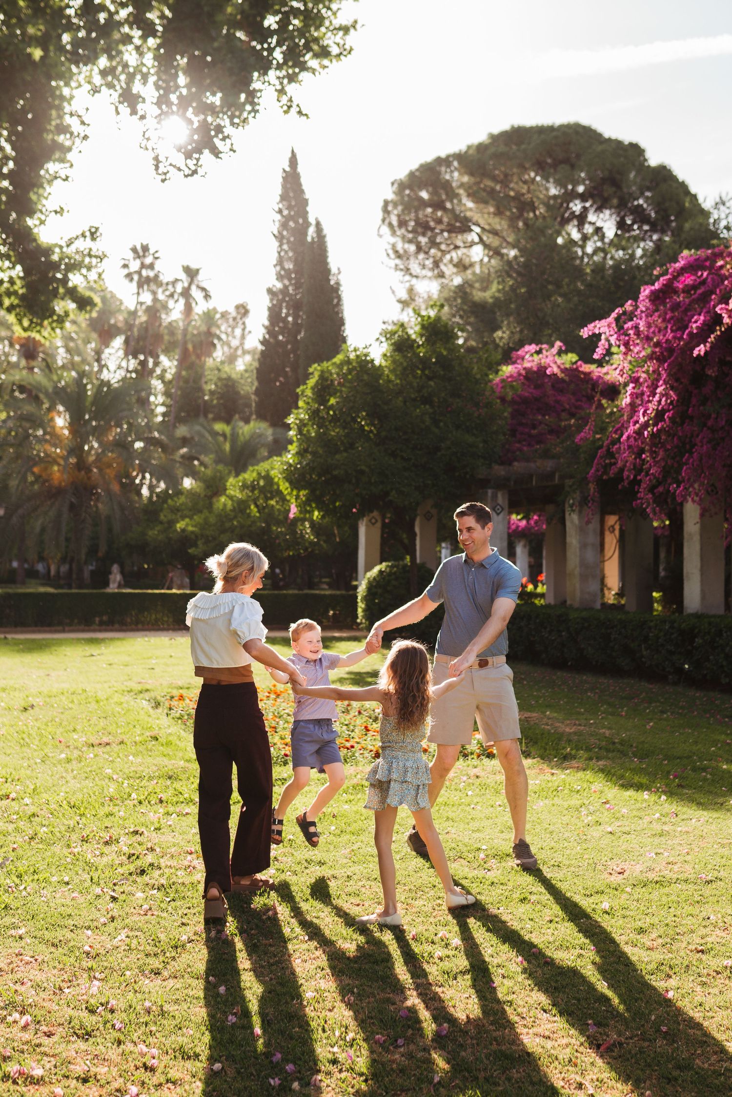 Family of four joyfully playing ring-around-the-rosie in a sunny park with colorful flowering trees.