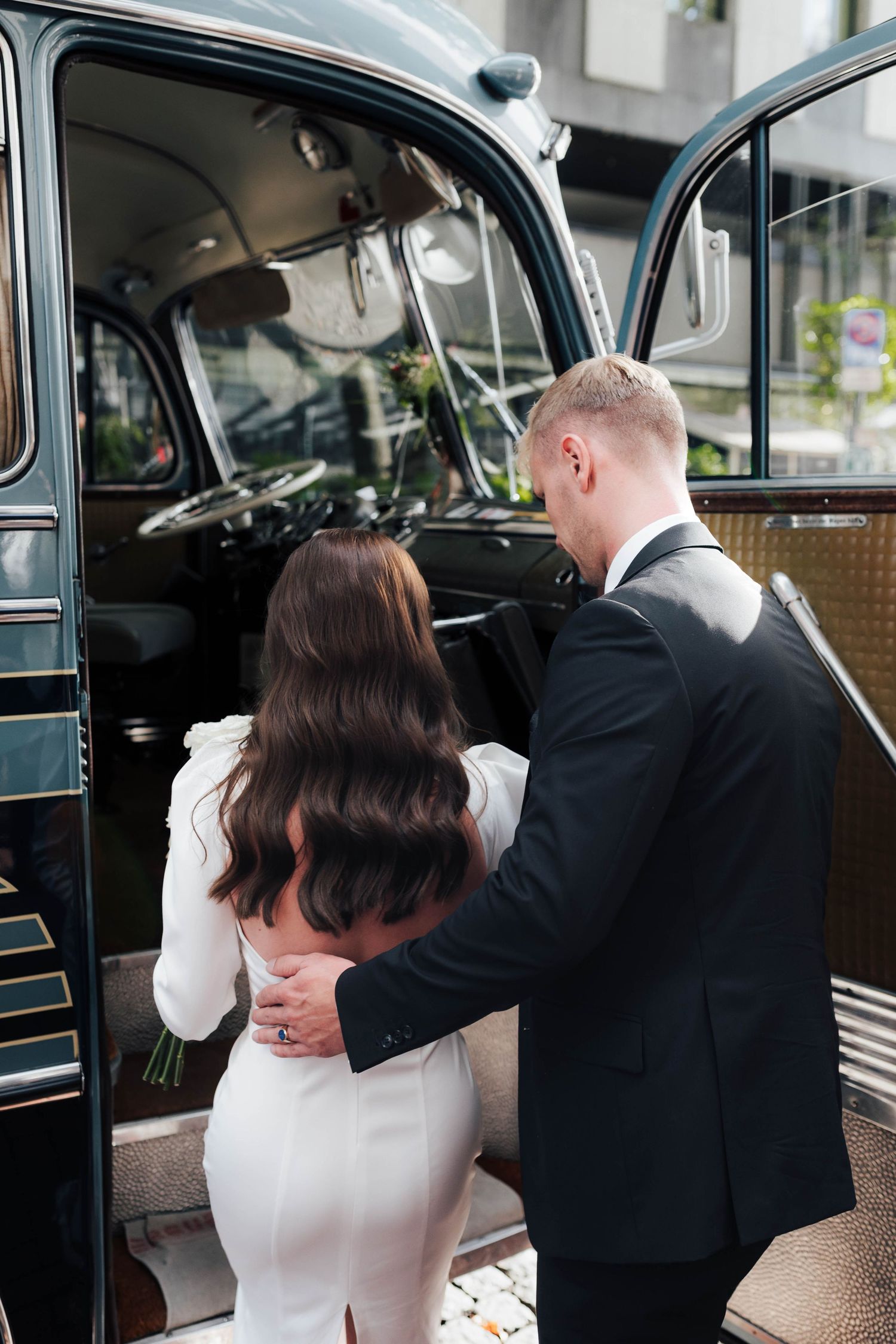A groom in a black suit helps his bride in a white dress into a classic black London taxi cab on their wedding day.