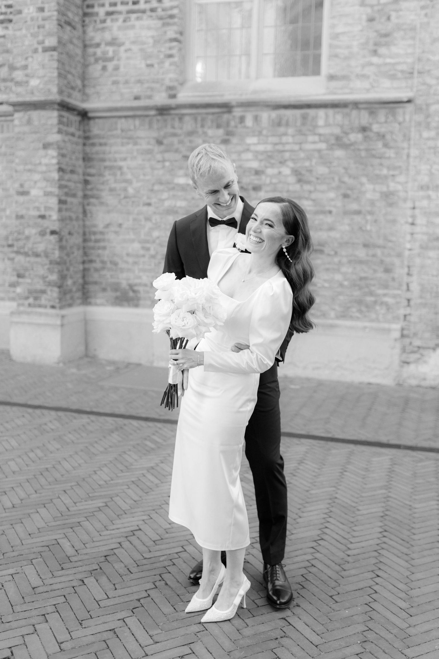 Black and white photo of a smiling bride in white midi dress holding bouquet, embraced by groom in tuxedo on brick street.