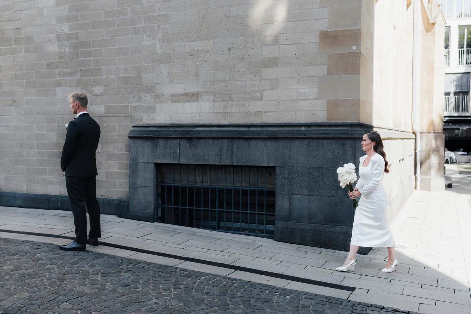 Bride in white dress holding bouquet approaches groom in black suit during outdoor first look wedding moment.