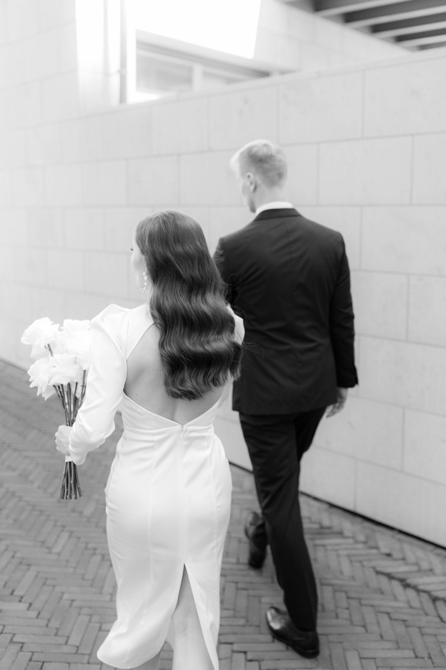 Black and white photo of bride and groom walking away, bride holding white bouquet in elegant backless gown.